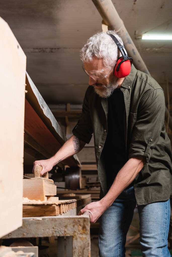 bearded carpenter in goggles polishing board in sander machine - 写真・画像