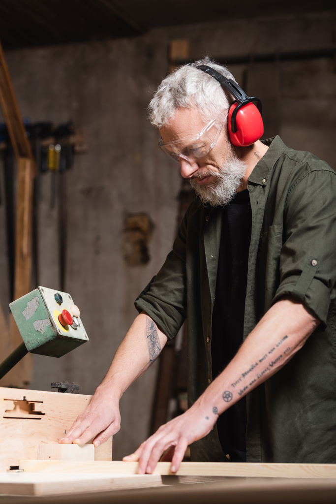 Sakallı marangoz koruyucu kulaklıklar takıyor. Jointer makinesinde çalışıyor.  - Fotoğraf, Görsel