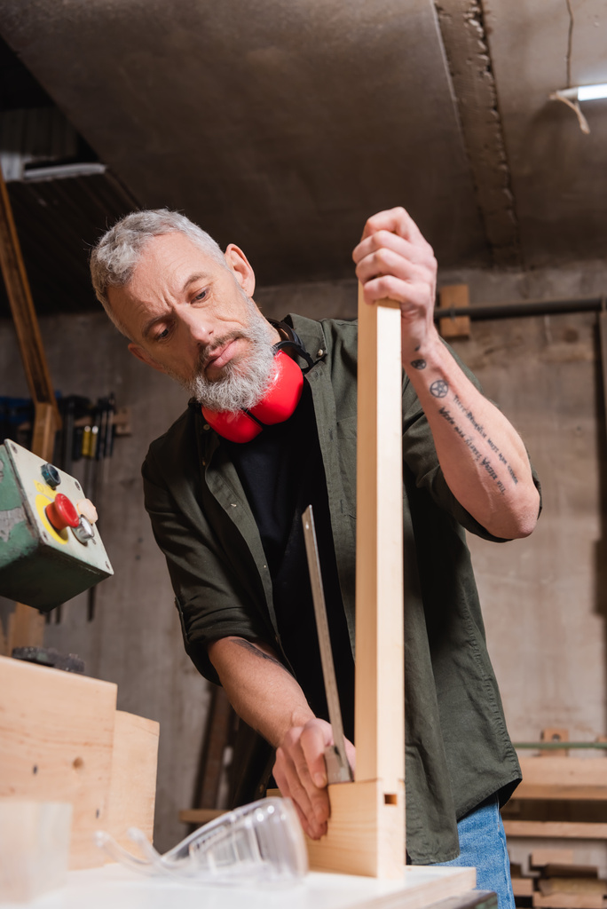 getatoeëerde timmerman controleren houten plank in houtwerk studio - Foto, afbeelding