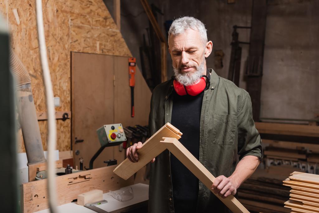 ώριμος γενειοφόρος σχεδιαστής επίπλων που κρατά ξύλινες σανίδες στο στούντιο ξυλουργικής - Φωτογραφία, εικόνα