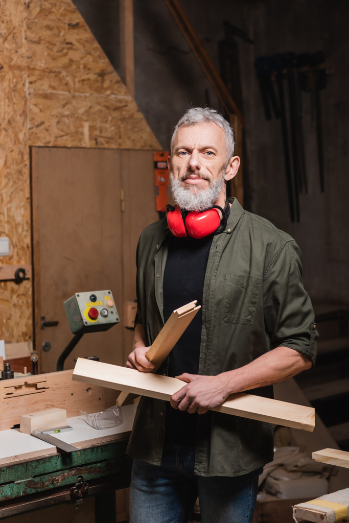 charpentier barbu tenant des détails en bois et regardant la caméra dans l'atelier - Photo, image