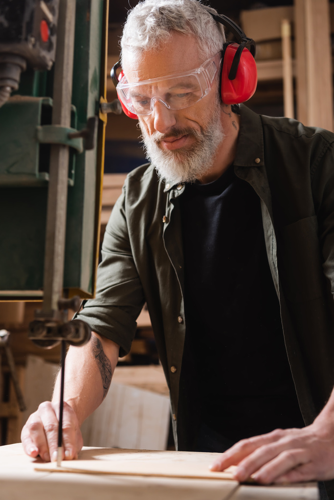 bearded carpenter in goggles cutting timber with band saw - Photo, Image
