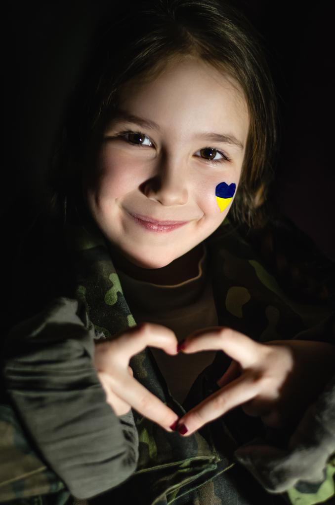 patriotic kid with painted ukrainian flag on cheek showing heart with hands and smiling isolated on black  - Photo, Image