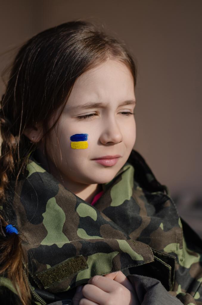 child with painted ukrainian flag on cheek and camouflage jacket - Photo, Image