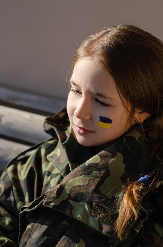 Сумна дитина з намальованим україніанським прапором на щоці та куртці з камуфляжу. - Фото, зображення
