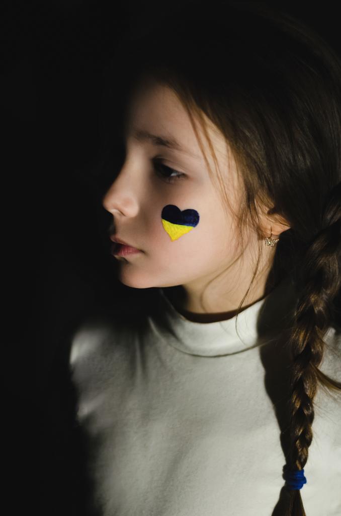 黒で隔絶された頬にウクライナの旗を描いたフラストレーションのある子供  - 写真・画像