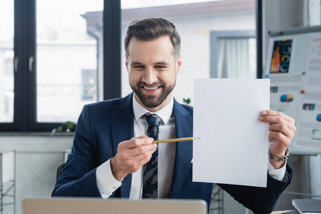 glimlachende econoom wijst met potlood naar leeg papier op kantoor - Foto, afbeelding