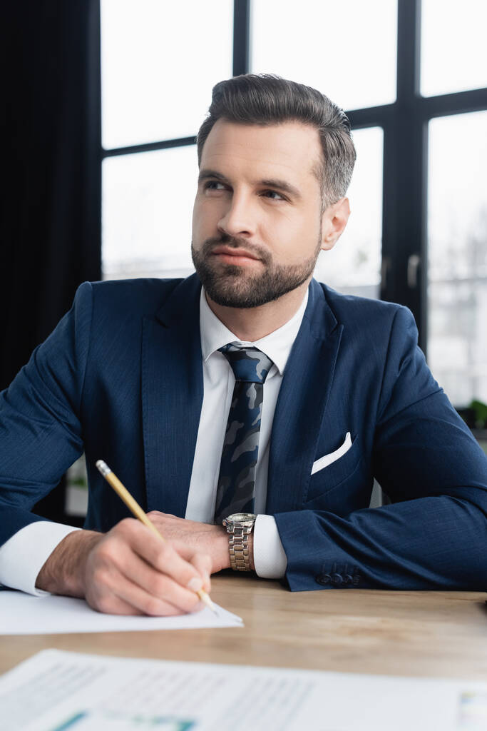economista reflexivo en chaqueta y corbata mirando hacia otro lado mientras escribe en la oficina - Foto, Imagen