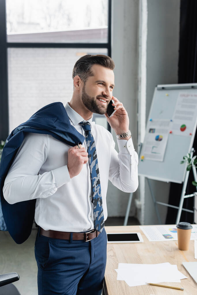 šťastný ekonom drží sako, zatímco mluví na smartphone v rozmazané kanceláři - Fotografie, Obrázek