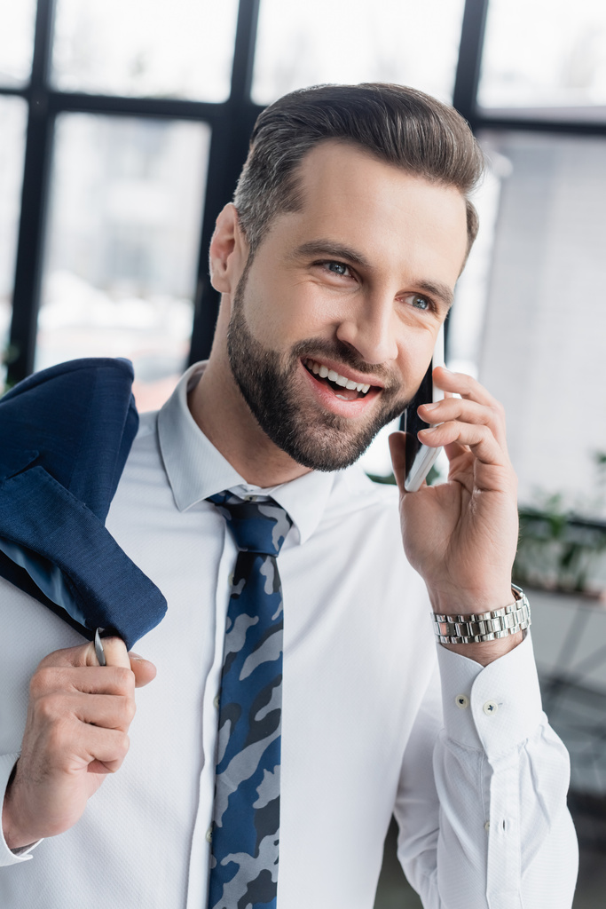 alegre hombre de negocios hablando en el teléfono móvil mientras sostiene la chaqueta en la oficina - Foto, imagen