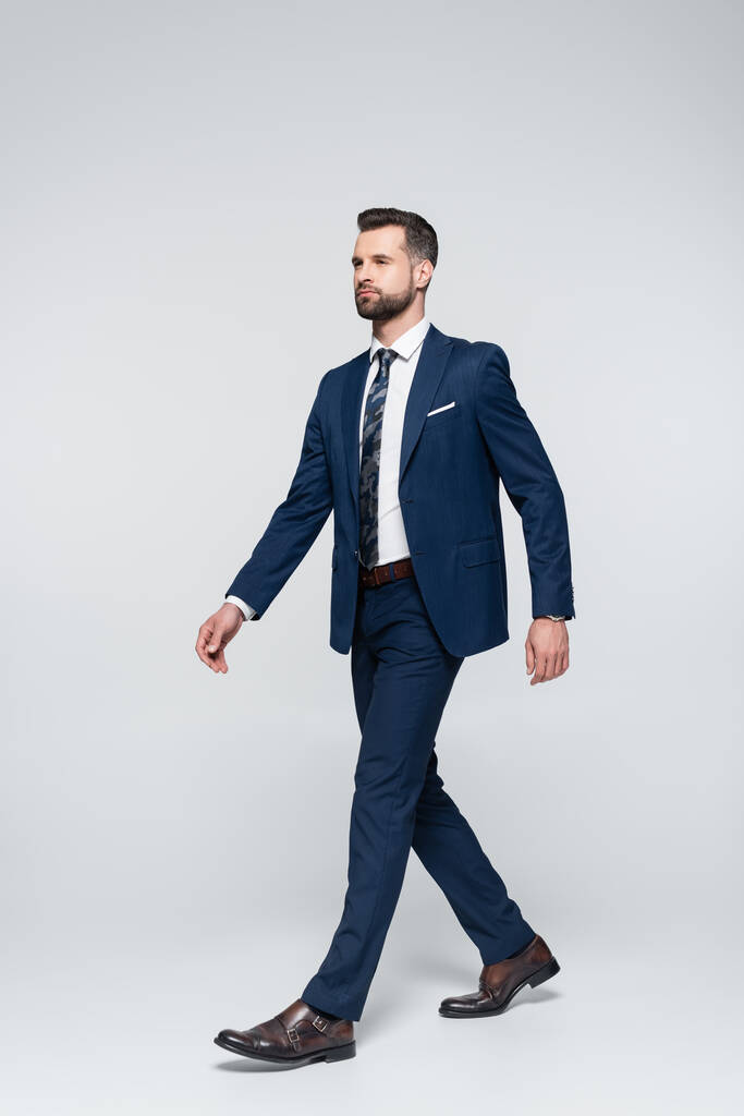 full length view of confident businessman in blue suit walking on grey - Фото, изображение