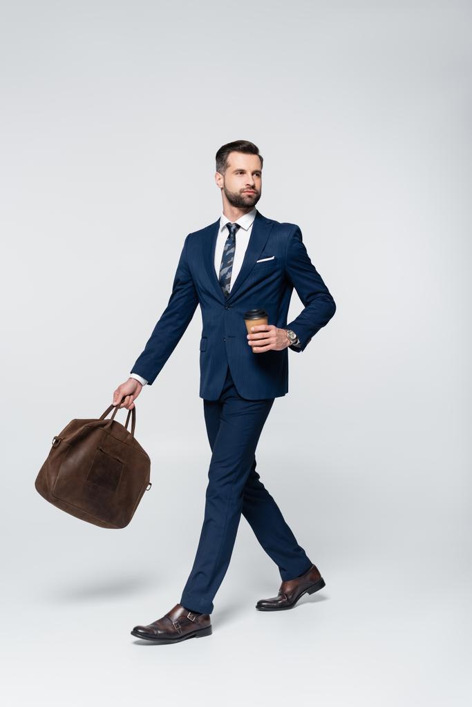 Mavi takım elbiseli ekonomist kahveyle yürüyor ve gri üzerine deri evrak çantası koyuyor. - Fotoğraf, Görsel