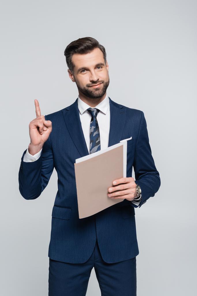 positive businessman holding folder and showing idea gesture isolated on grey - Фото, изображение