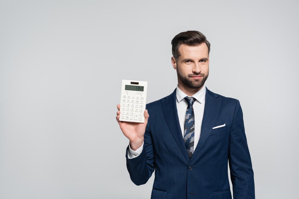 economista morena mostrando calculadora e sorrindo para a câmera isolada em cinza - Foto, Imagem