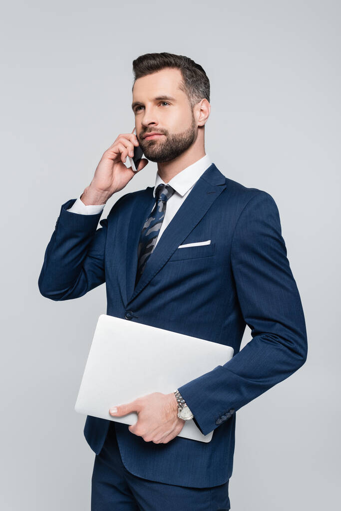 hombre de negocios barbudo con portátil hablando en el teléfono móvil aislado en gris - Foto, imagen