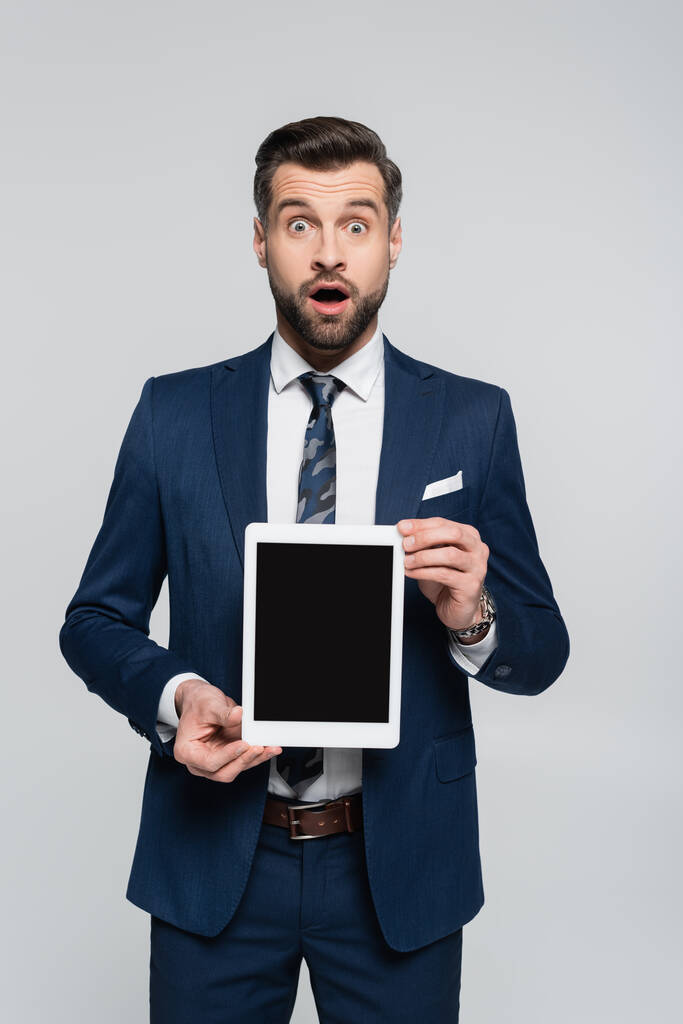 užaslý ekonom ukazující digitální tablet s prázdnou obrazovkou na kameru izolovanou na šedé - Fotografie, Obrázek