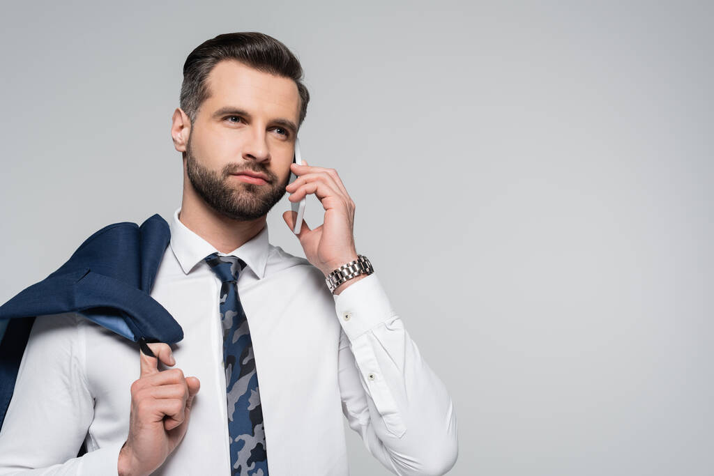 brunette businessman holding blazer, talking on smartphone and looking away isolated on grey - Photo, Image
