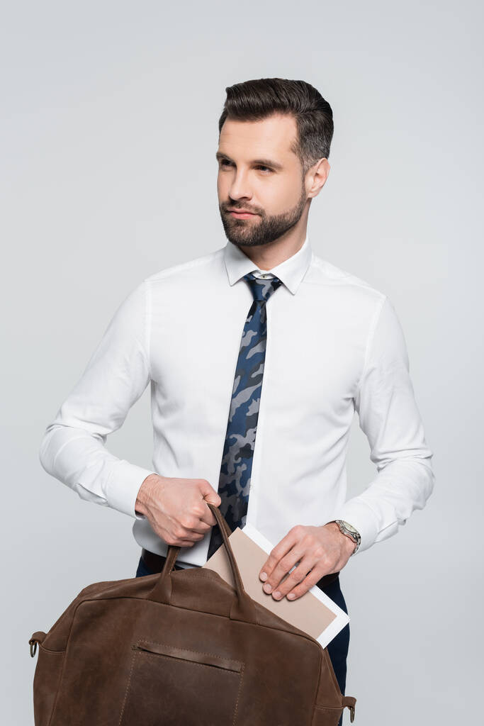 επιχειρηματίας σε λευκό πουκάμισο κρατώντας χαρτοφύλακα και φάκελο, ενώ κοιτάζοντας μακριά απομονώνονται σε γκρι - Φωτογραφία, εικόνα