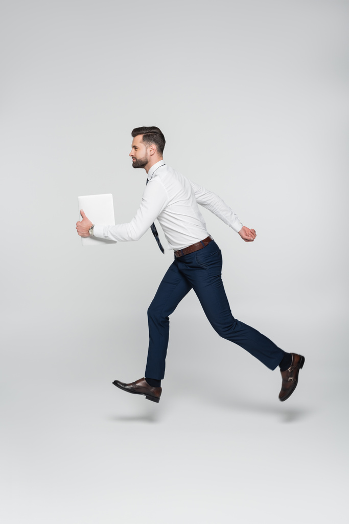 vista lateral de hombre de negocios en camisa blanca corriendo con tableta digital en gris - Foto, Imagen