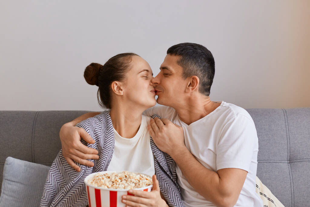 Indoor shot van liefdevolle romantische vrouw en echtgenoot zitten op de bank met pop-maïs en afstandsbediening, kijken naar romantische film, zoenen terwijl, uiten van liefde en zachte. - Foto, afbeelding