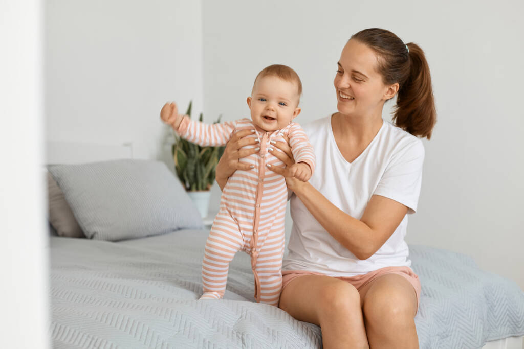 Indoor shot of woman with dark hair and ponytail wearing white t shirt and shorts sitting on bed with her baby daughter, kid wearing striped sleeper, mother playing with child. - Photo, Image