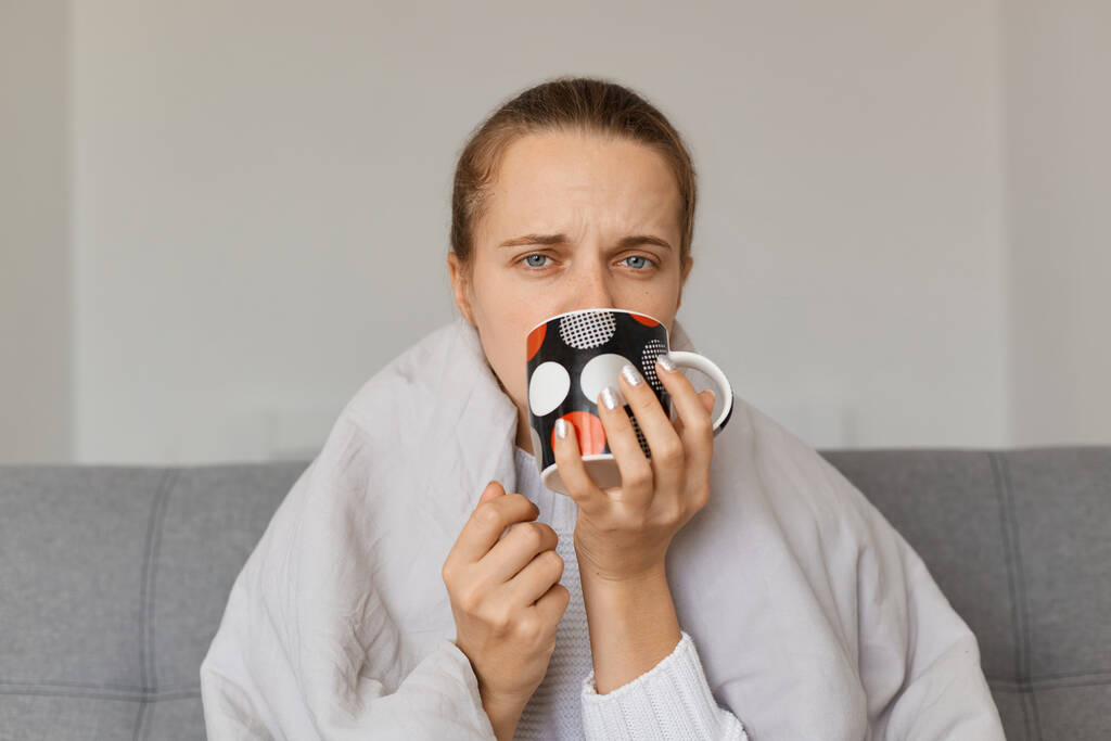Retrato de una joven adulta malsana envuelta en una manta sentada en un sofá y bebiendo té caliente, resfriada, con síntomas de gripe o grippe. - Foto, imagen
