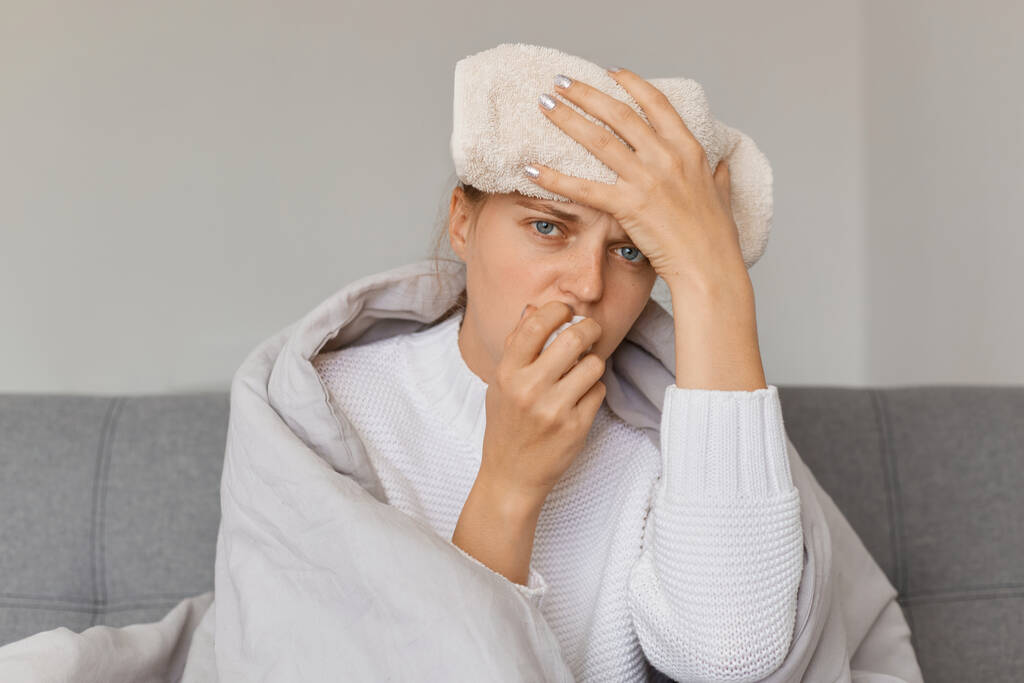 Горизонтальный снимок белой больной женщины, завернутой в одеяло, сидящей на диване с мокрым полотенцем на лбу, смотрящей в камеру с расстроенным лицом, страдающей головной болью, насморком и высокой температурой. - Фото, изображение