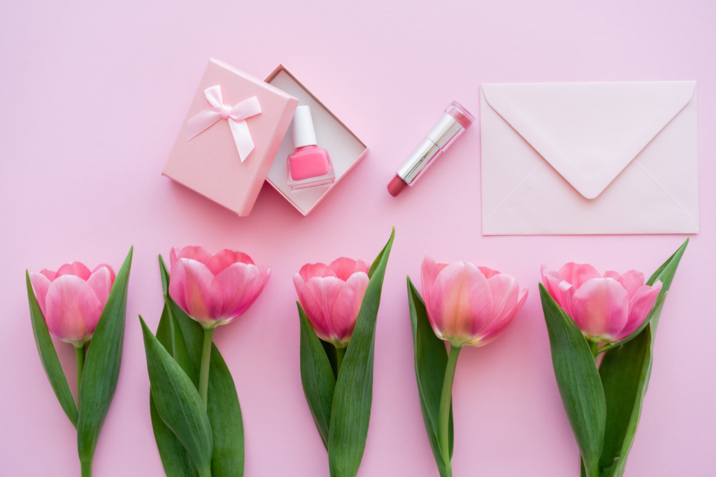 vue du haut de la rangée avec des tulipes en fleurs près de la boîte cadeau avec vernis à ongles près du rouge à lèvres et enveloppe sur rose - Photo, image