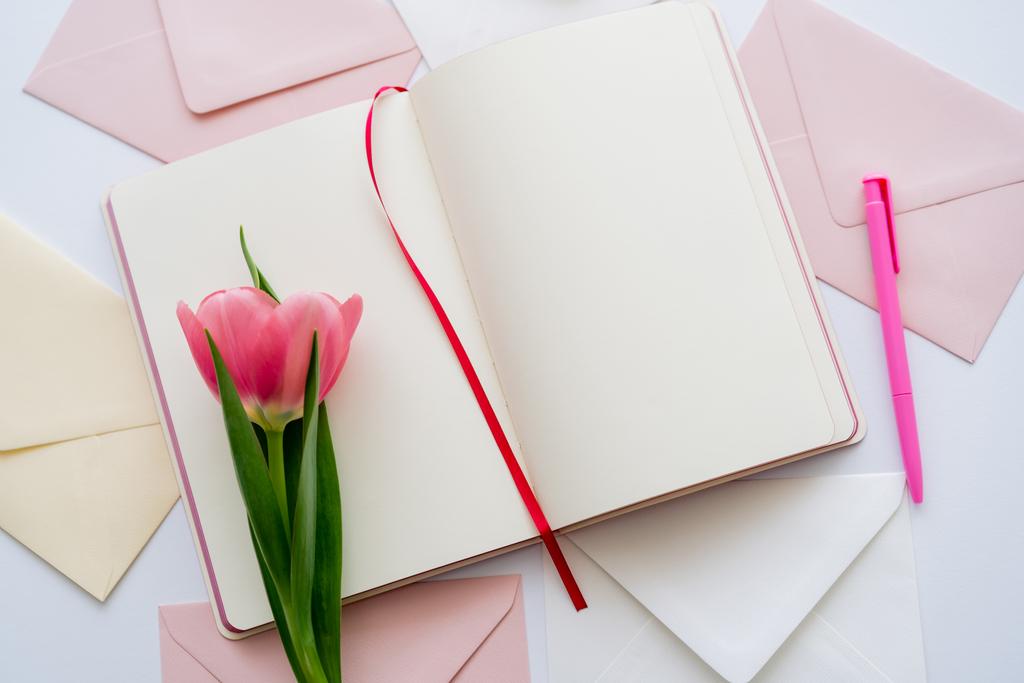 bovenaanzicht van roze tulp op blanco notitieboekje bij enveloppen op wit - Foto, afbeelding