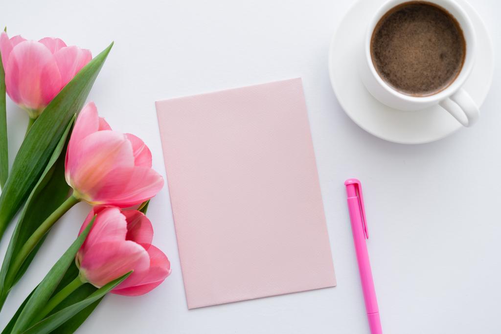 vue du dessus de tasse de café près de tulipes roses, papier et stylo sur blanc  - Photo, image