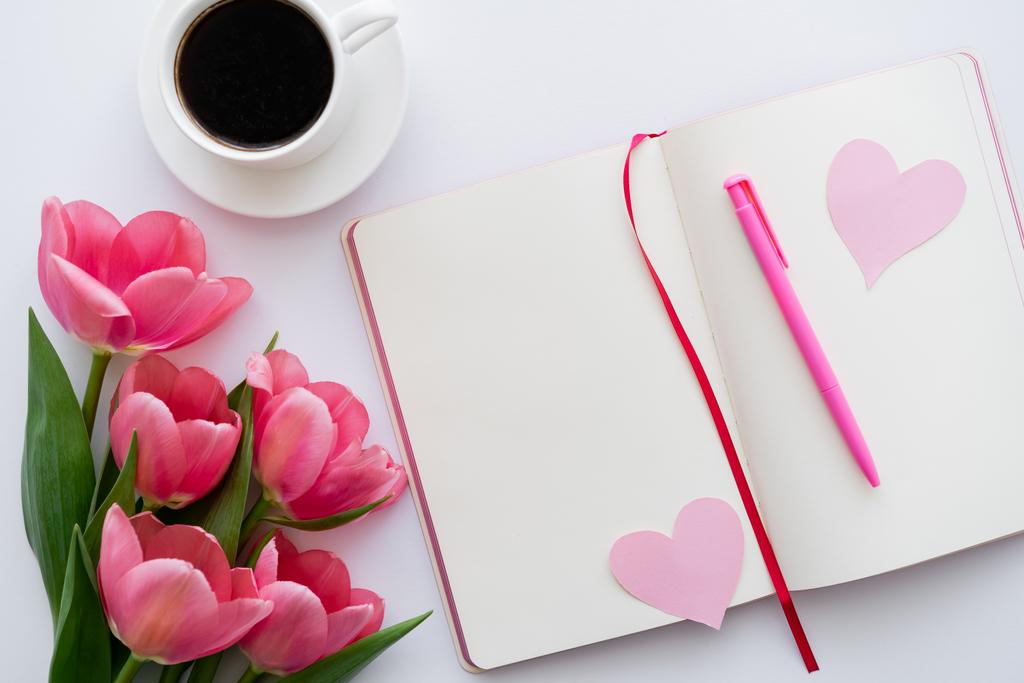 top view of tulips near notebook with paper hearts and cup of coffee on white - Photo, Image