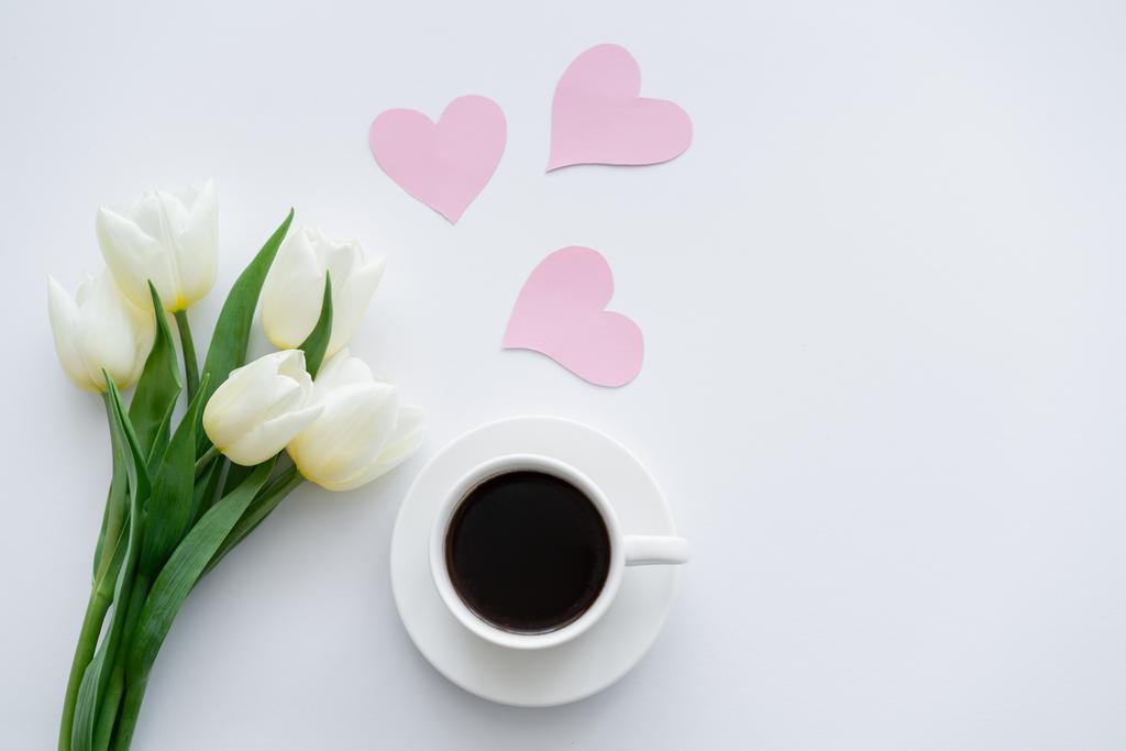 bovenaanzicht van tulpen in de buurt van kopje met koffie op schotel in de buurt van papieren harten op wit - Foto, afbeelding