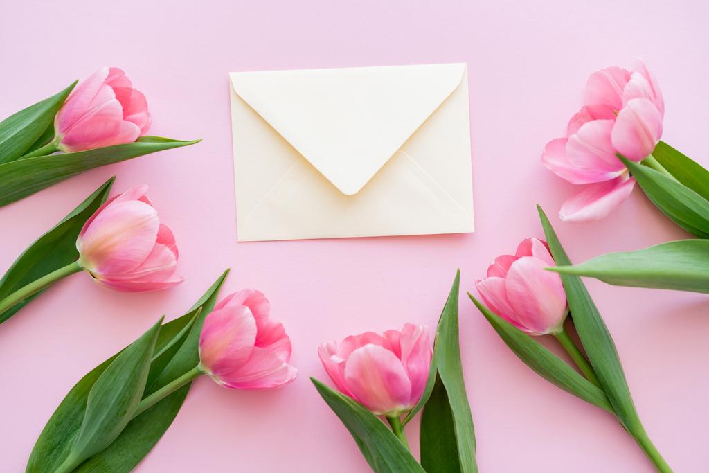 flat lay of blossoming tulips with green leaves near envelope on pink, mothers day concept  - Photo, Image