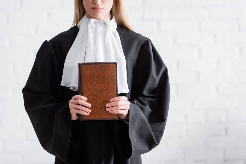 partial view of prosecutor in black mantle holding book near white wall - Foto, immagini
