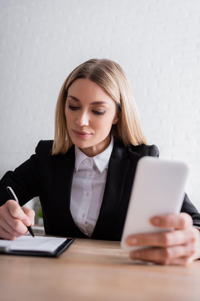 lawyer holding smartphone and writing in notebook while working in office - Photo, Image