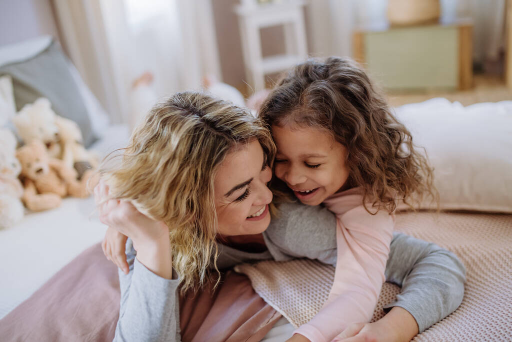 Ευτυχισμένη μητέρα με την κορούλα της να αγκαλιάζεται και να διασκεδάζει μαζί στο κρεβάτι στο σπίτι. - Φωτογραφία, εικόνα