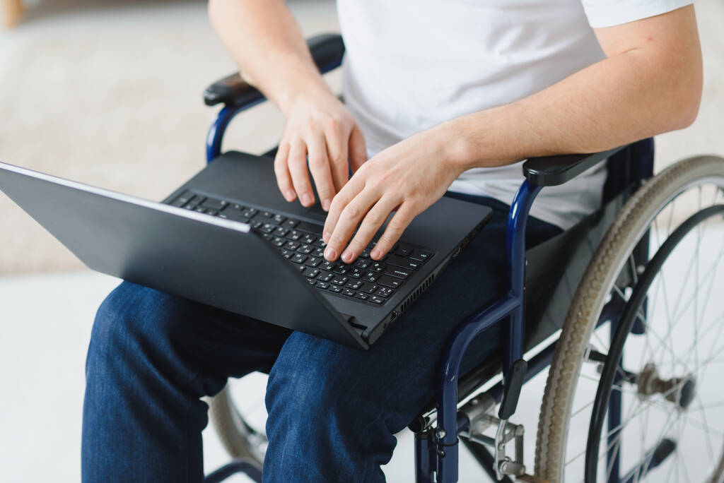 Πορτρέτο του χαμογελαστού ανάπηρου άνδρα που κάθεται σε αναπηρική καρέκλα και εργάζεται σε φορητό υπολογιστή από το σπίτι. Νέος εργαζόμενος με ειδικές ανάγκες. Freelancer και άτομα με ειδικές ανάγκες έννοια. - Φωτογραφία, εικόνα