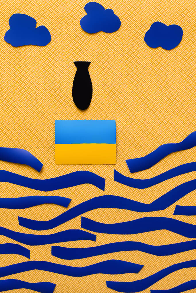 Vista dall'alto della bomba di carta vicino alla bandiera ucraina, nuvole e mare di cartone su sfondo giallo strutturato  - Foto, immagini