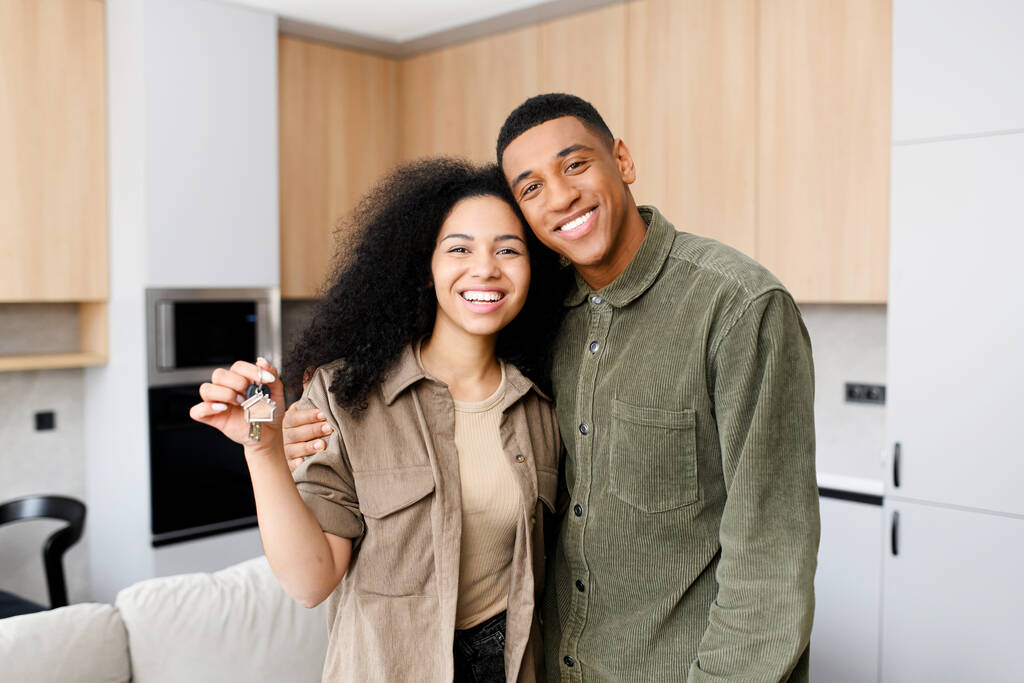 Excited multiracial couple in love move in a new apartment. Cheerful woman in embrace of her guy holding the keys - Photo, Image