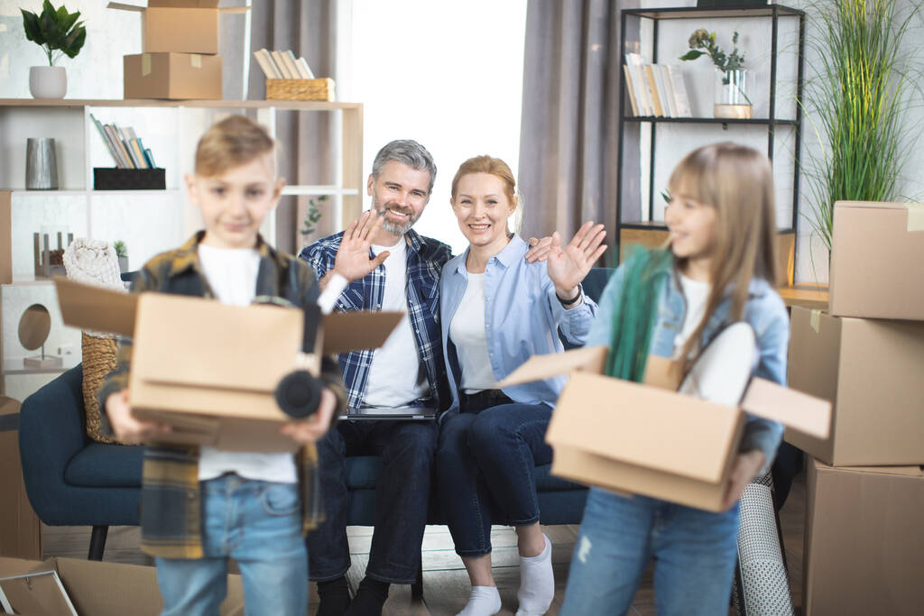 Enfants souriants avec des boîtes mobiles posant dans un nouvel appartement avec des parents derrière - Photo, image