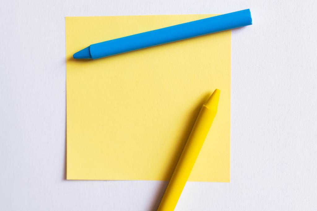 Beyaz üzerine izole edilmiş kağıdın yanındaki mavi ve sarı boya kalemlerinin üst görünümü  - Fotoğraf, Görsel