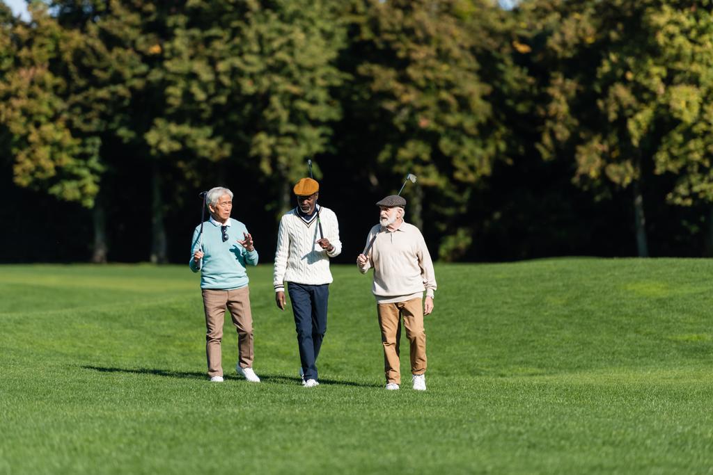 amigos seniores inter-raciais andando com tacos de golfe em campo - Foto, Imagem