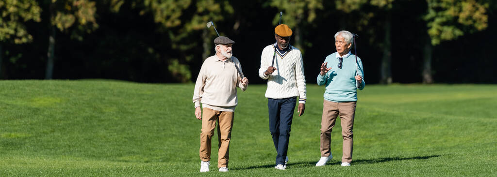interracial senior friends walking with golf clubs on field, banner - 写真・画像