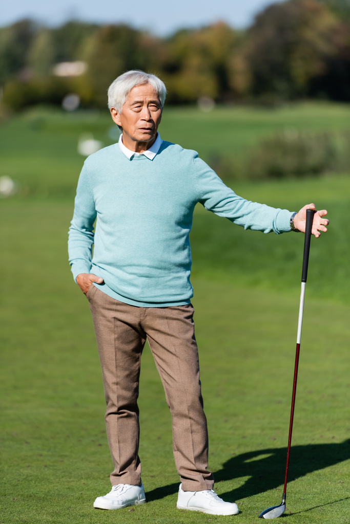 азіатський старший чоловік стоїть з рукою в кишені і гольф-клубі на зеленому полі
 - Фото, зображення