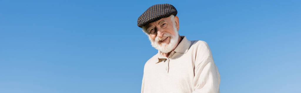 šťastný starší muž v ploché čepici při pohledu na kameru proti modré obloze, prapor - Fotografie, Obrázek