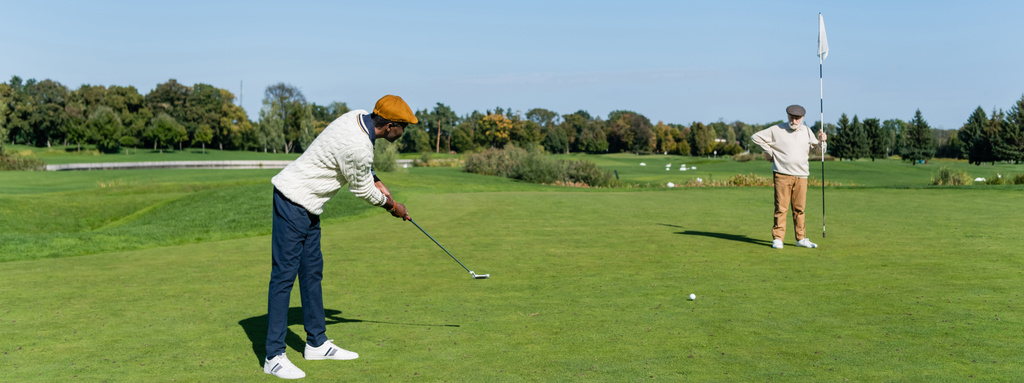 homem sênior em cap plana segurando bandeira vara enquanto amigo afro-americano jogando golfe, banner - Foto, Imagem