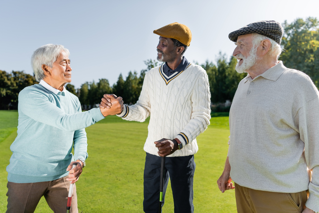 senior asian and african american men shaking hands while holding golf clubs near cheerful friend   - Фото, изображение