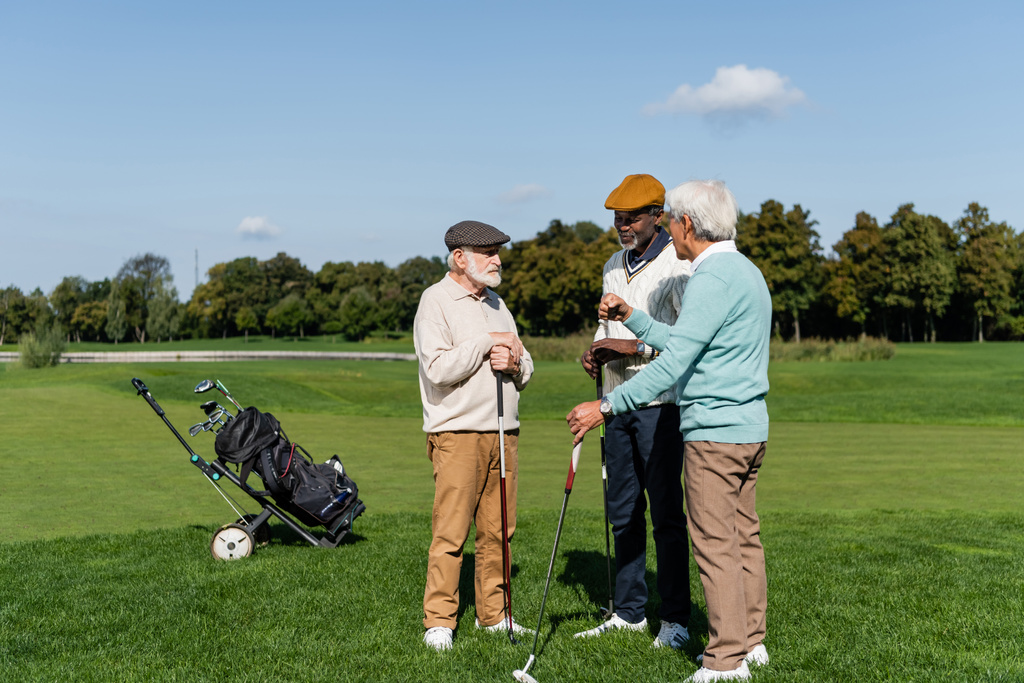 amigos multiétnicos seniores segurando tacos de golfe e falando em campo  - Foto, Imagem