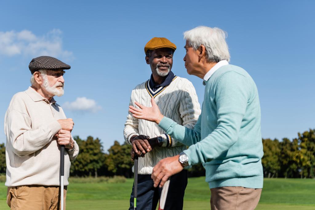 старшие межрасовые друзья, держащие клюшки для гольфа и разговаривающие на поле  - Фото, изображение