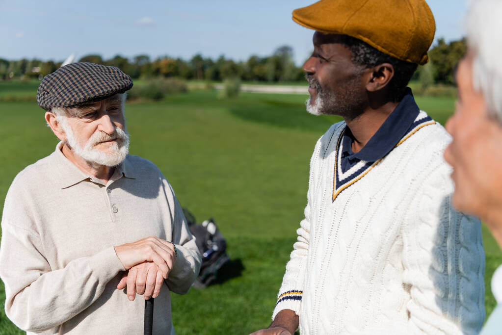 amigos multiétnicos senior mirando al hombre en gorra plana en el campo de golf  - Foto, imagen